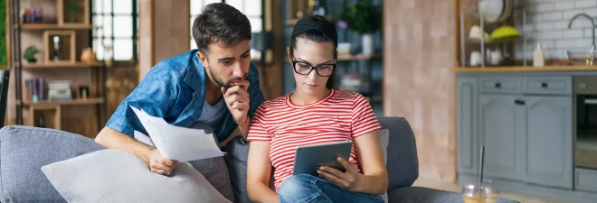 Couple calculating bills at home using tablet and calculator. Young couple working on computer while calculating finances sitting on couch. Young man with wife at home analyzing their finance with documents