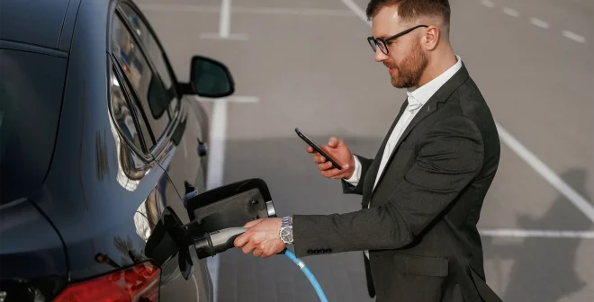 Charging the electric vehicle. Businessman in suit is near his black car outdoors