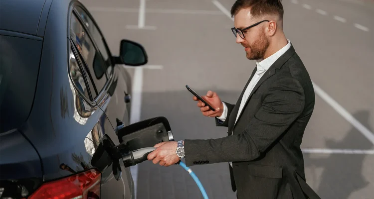 Charging the electric vehicle. Businessman in suit is near his black car outdoors